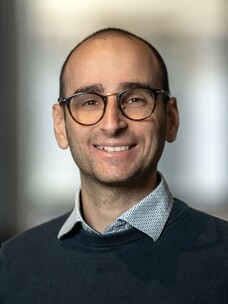 Herr Disante mit Brille, leichtem Lächeln und kahlem Kopf vor neutralem Hintergrund.